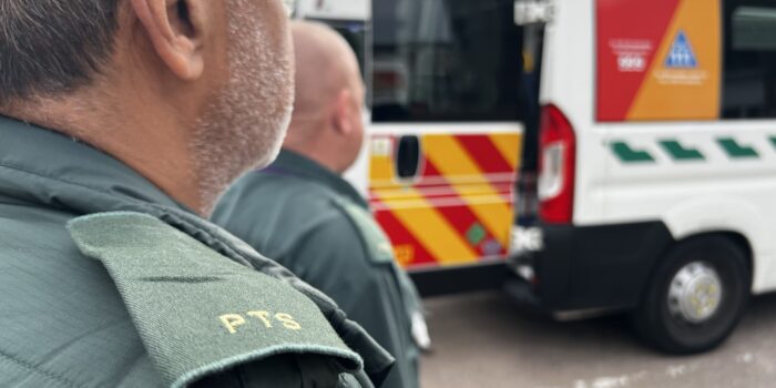 Photo over the shoulders of two male patient transport service staff standing at back of ambulance with door open