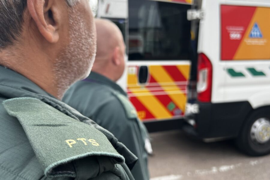 Photo over the shoulders of two male patient transport service staff standing at back of ambulance with door open