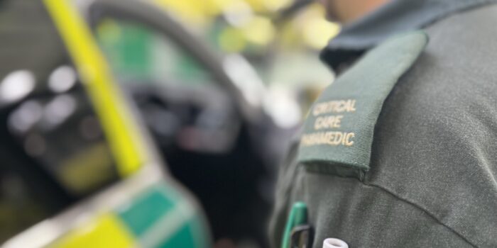 Man in green WMAS uniform walking towards open ambulance response car. Focus of photo is over left shoulder showing 'critical care paramedic' epaulette