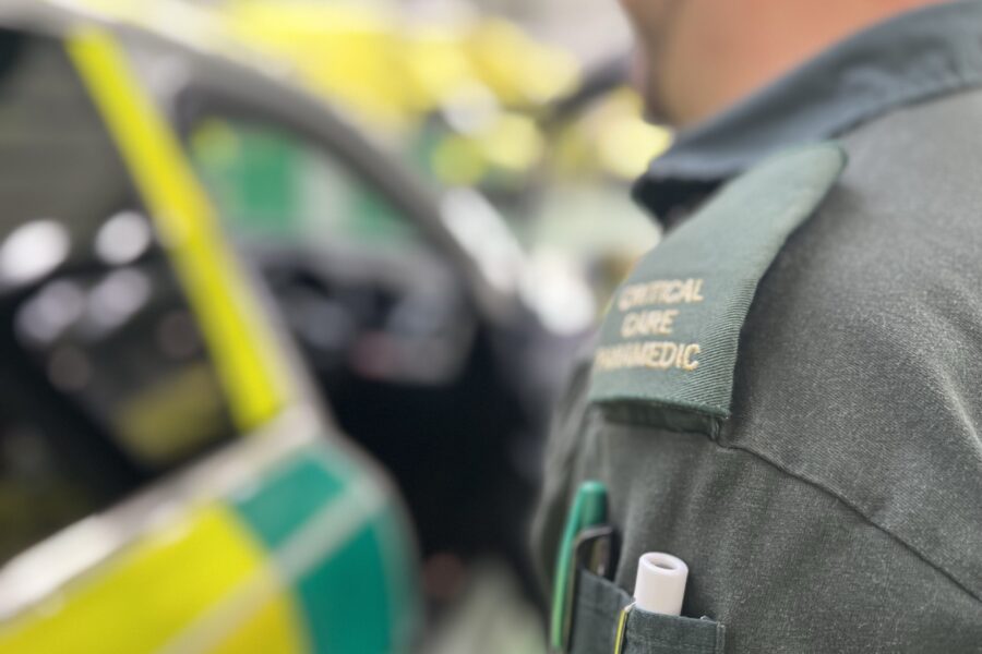 Man in green WMAS uniform walking towards open ambulance response car. Focus of photo is over left shoulder showing 'critical care paramedic' epaulette