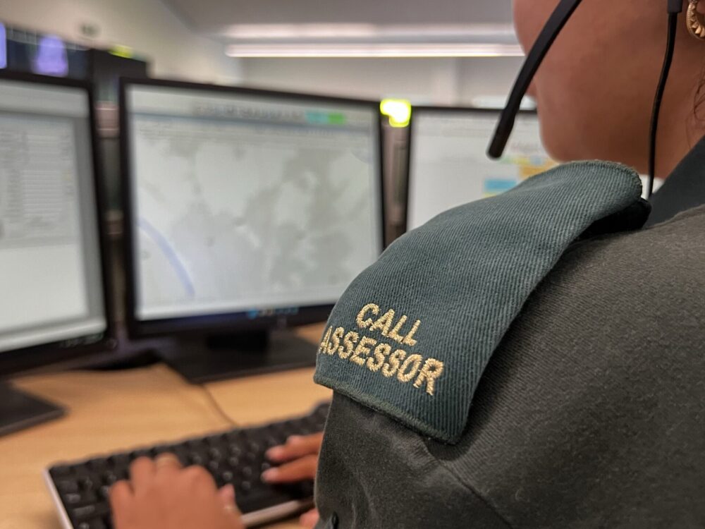 Over the shoulder photo of Call Assessor wearing epaulettes and green uniform with headset on and typing on keyboard whilst looking at computer screens.