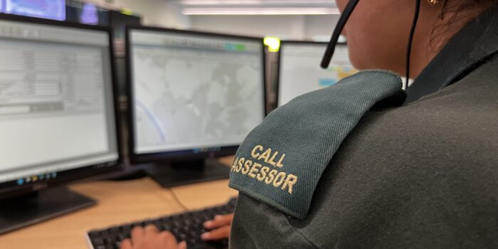Over the shoulder photo of Call Assessor wearing epaulettes and green uniform with headset on and typing on keyboard whilst looking at computer screens.