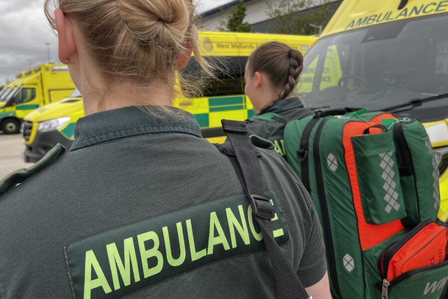 Two female ambulance staff wearing kit bags on back, walking away from camera with ambulances lined in in background