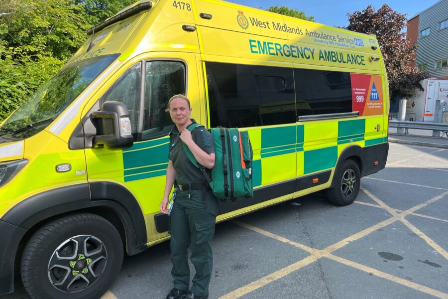 Paramedic Hannah standing at side of ambulance with uniform and kit bag on back