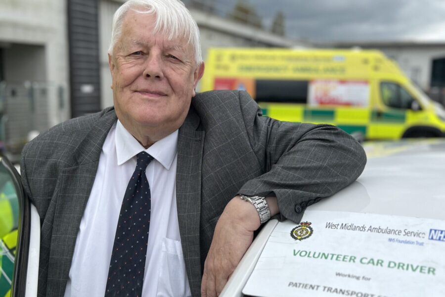 Volunteer car driver William Read smiling at camera whilst leaning on roof of car with drivers door open. Ambulance in background and sign on windscreen of car saying 'volunteer car driver'