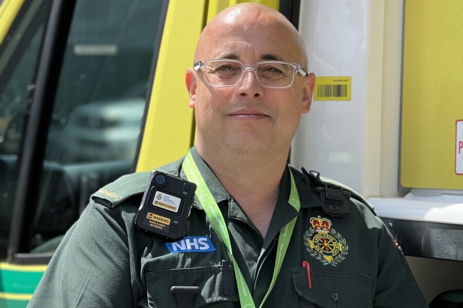 Mental Health paramedic Tony Sutton in uniform, standing next to ambulance with side door open and leaning on shelf inside with one arm. Smiling at camera.