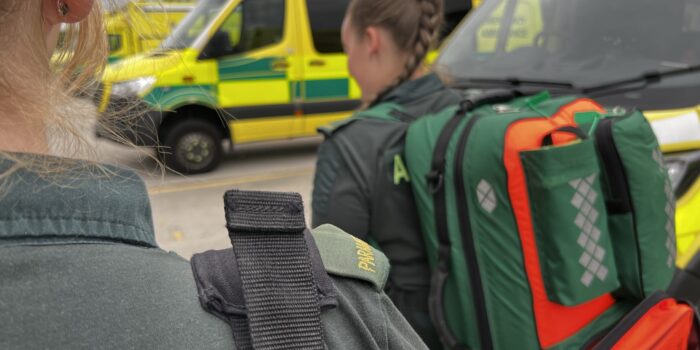 Two female parmedics with kit bags on back walking away from camera past row of parked ambulances