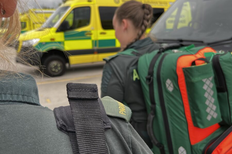 Two female parmedics with kit bags on back walking away from camera past row of parked ambulances