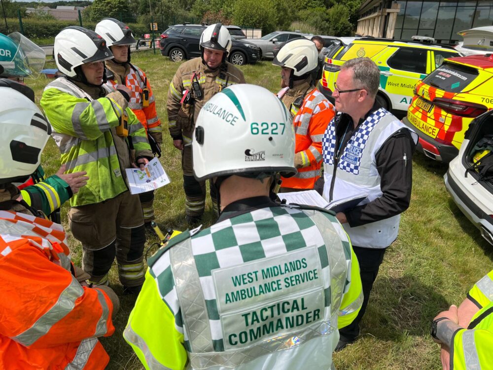 Group of emergency service staff from fire, police and ambulance wearing full uniform and helmets in a circle talking with each other at an incident
