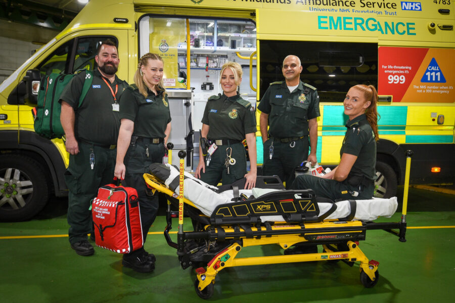 Five ambulance staff in green uniform smiling at camera and standing in front of ambulance with door open with stretcher in front of them.