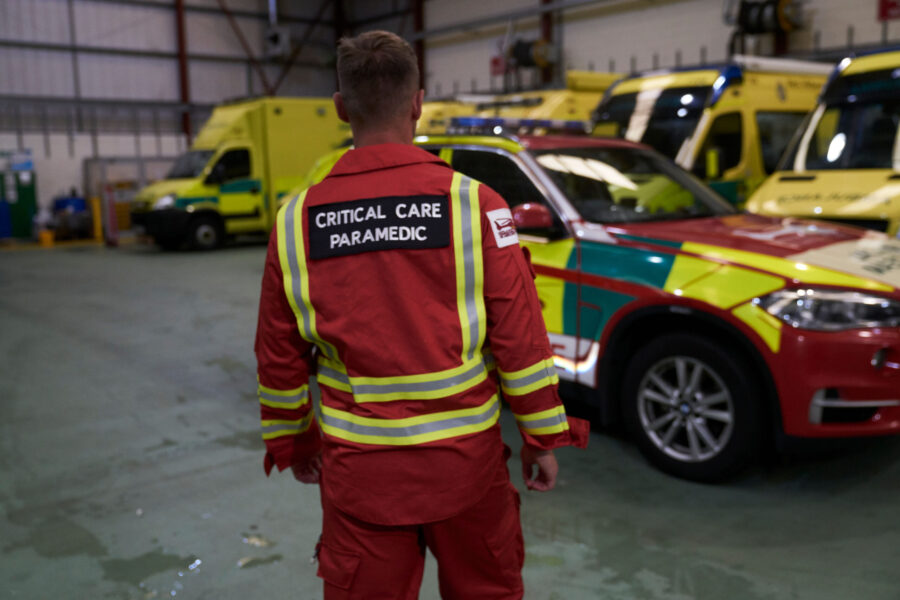 Ambulance Code Red: Critical Care Paramedic in red jumpsuit uniform with back to camera walking towards red critical care car parked in front of ambulance line up.