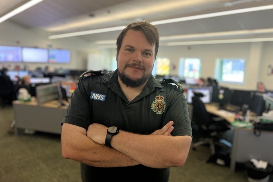 Control room dispatcher Aaron smiling at camera with arms folded whilst standing in control room. Desks and controllers in background blurred.