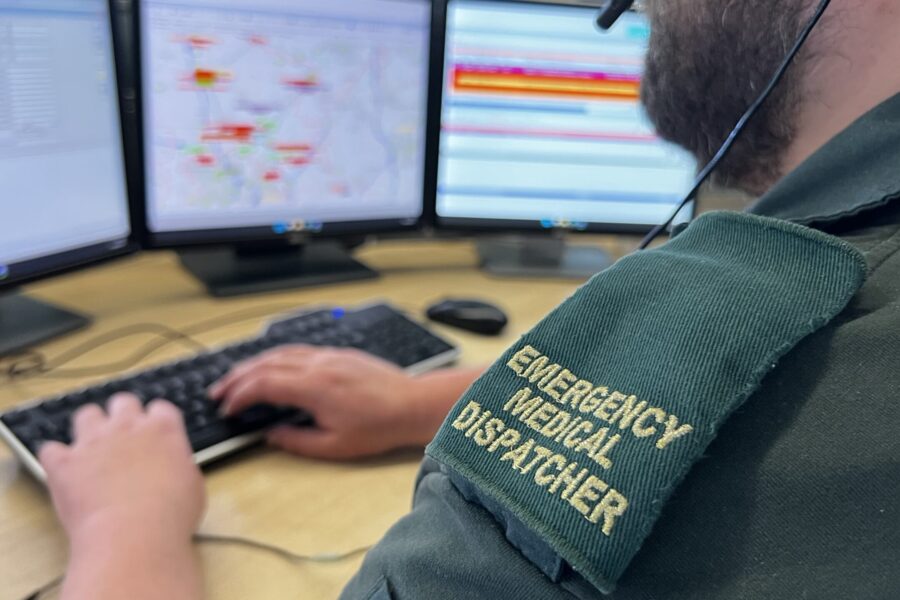 Male dispatcher with beard and green epaulettes on shoulder typing looking at bank of screens