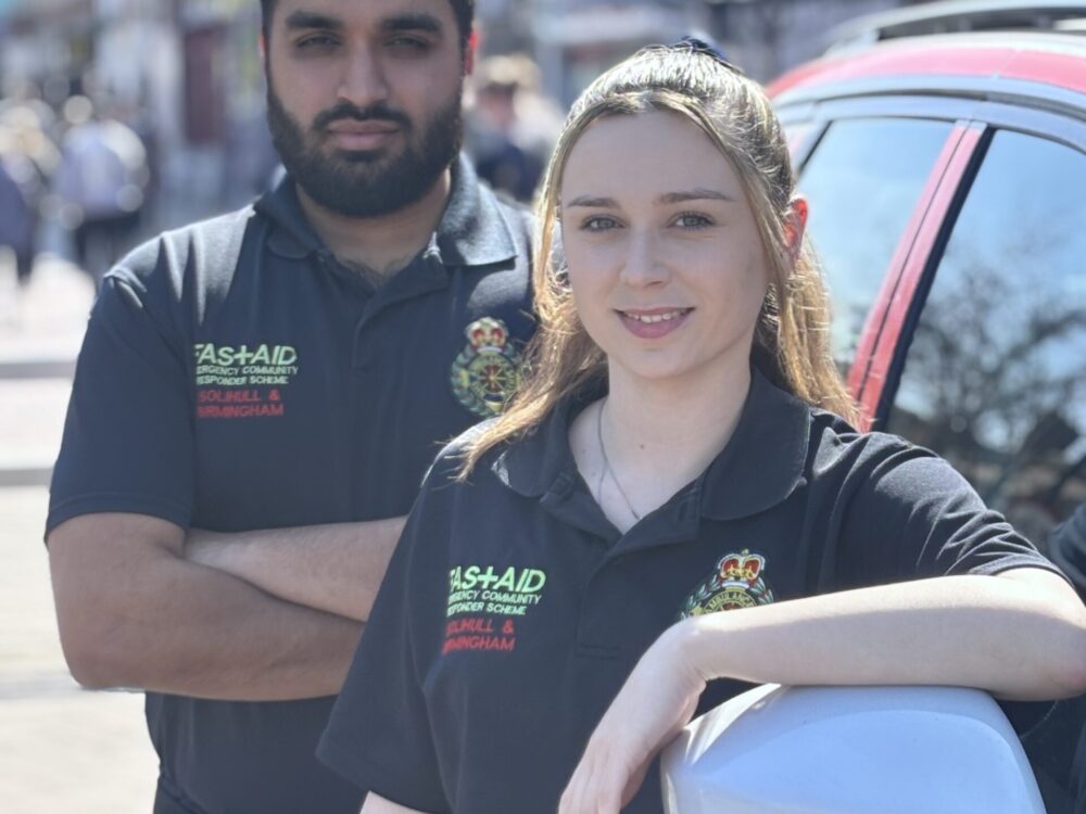 A male and female Community First Responder smiling at camera wearing blue CFR uniform, standing next to CFR vehicle in busy high streer