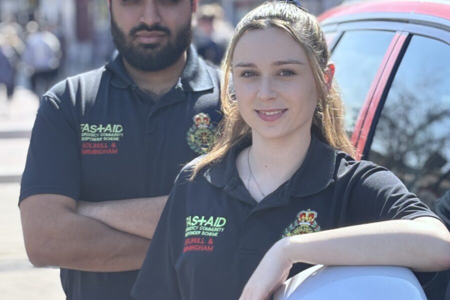 A male and female Community First Responder smiling at camera wearing blue CFR uniform, standing next to CFR vehicle in busy high streer